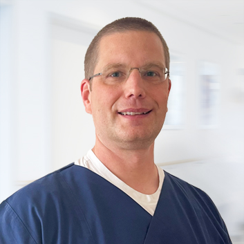 Porträtbild des Arztes Dr. med. Heimann vor einem hellem Hintergrund. Der Arzt trägt einen dunkelblauen Kasack.