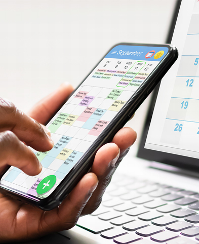 Eine Person hält ein Smartphone in der Hand – auf dem Display ist ein Kalender zu sehen. Im Hintergrund sieht man einen aufgeklappten Laptop auf einem Tisch – hier sieht man ebenfalls einen Kalender auf dem Bildschirm.