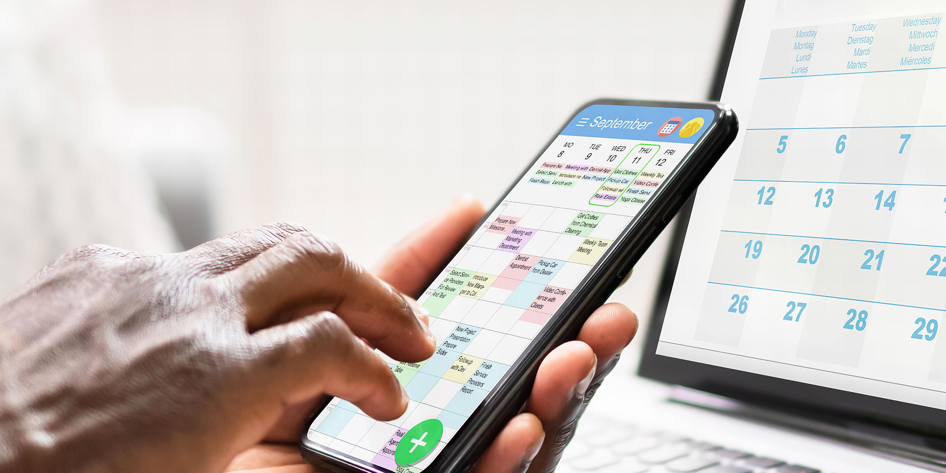 Eine Person hält ein Smartphone in der Hand – auf dem Display ist ein Kalender zu sehen. Im Hintergrund sieht man einen aufgeklappten Laptop auf einem Tisch – hier sieht man ebenfalls einen Kalender auf dem Bildschirm.