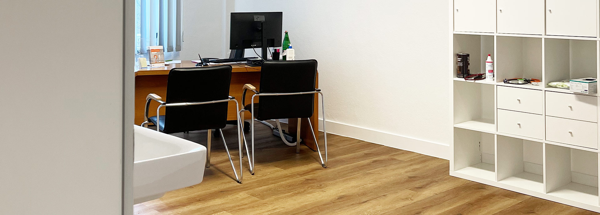 Blick in einen der Behandlungsräume der Praxis mit Schreibtisch, weißem Regal und einem Wandbild zum Thema 