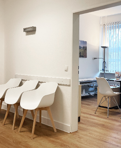 Blick vom Wartebereich der Praxis mit weißen Stühlen in einen modern gestalteten Behandlungsraum der Praxis.