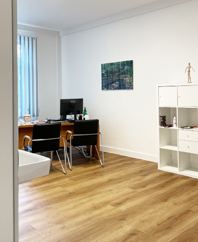 Blick in einen der Behandlungsräume der Praxis mit Schreibtisch, weißem Regal und einem Wandbild zum Thema 